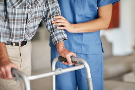 An image of a nurse supporting a man as he walks with the help of a walker.