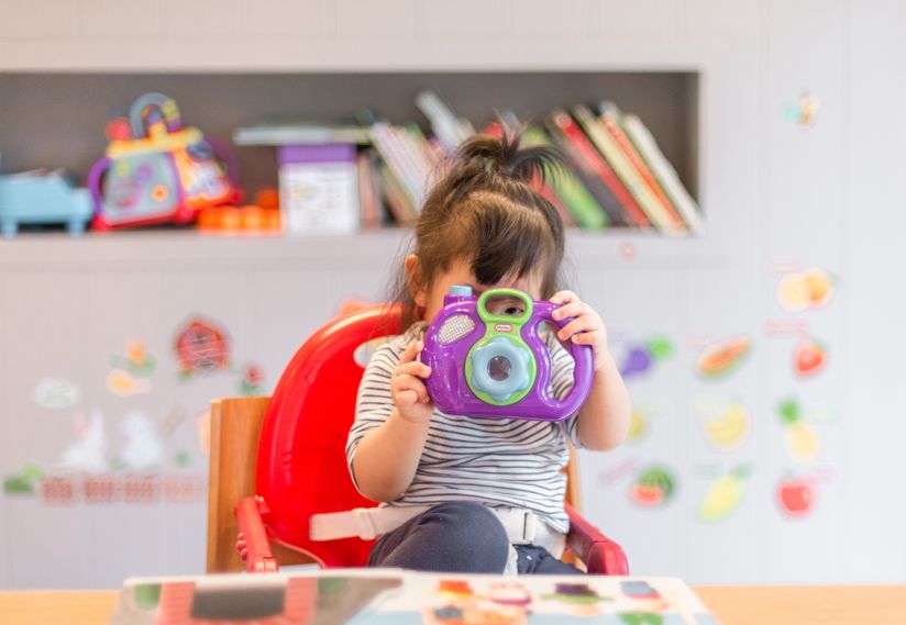 An adorable young girl sitting in a high chair, playfully grasping a camera while engaging with the surroundings. An adorable young girl sitting in a high chair, playfully grasping a camera while engaging with the surroundings.