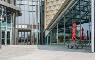 Modern office building exterior with glass windows, outdoor seating, and red umbrellas in a commercial space.